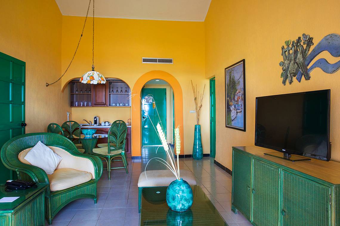 Innenansicht eines Wohnzimmers im Memories Jibacoa Resort mit gelben Wänden, grünen Möbeln und einer großen Fernsehwand.