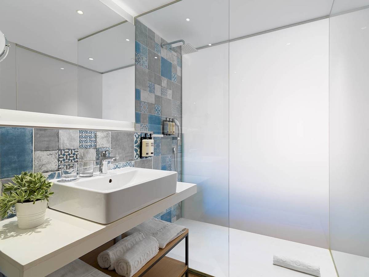 Badezimmer im H10 Casa del Mar Hotel mit blauen und grauen Fliesen, weißem Waschbecken und Glasdusche.