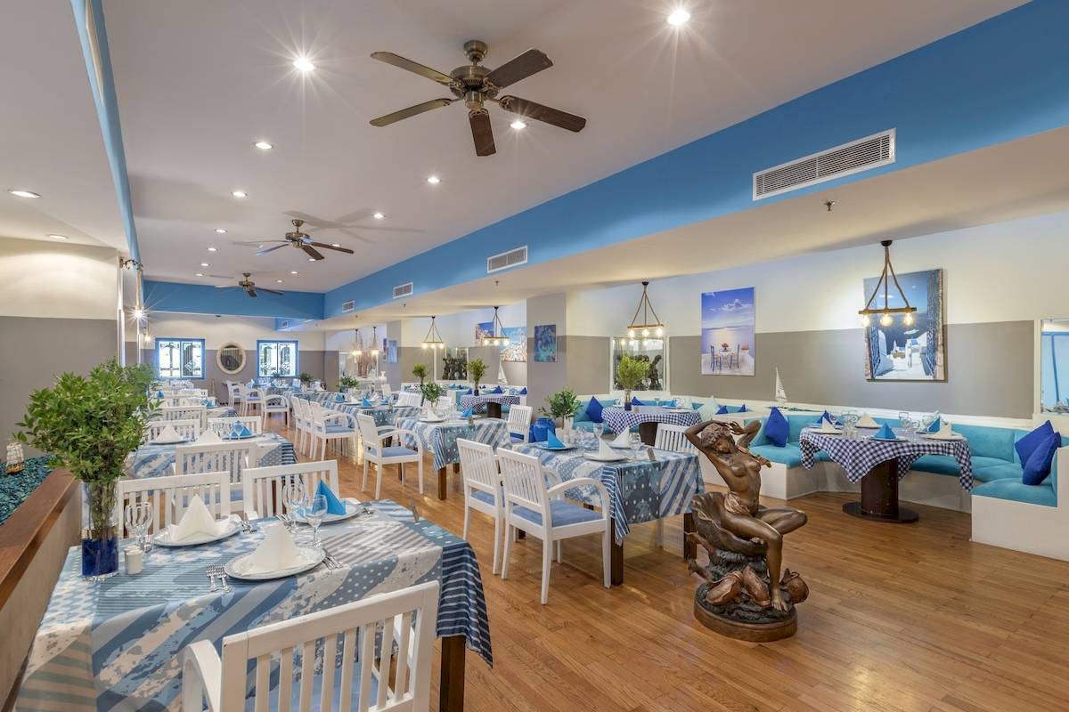 Innenbereich des Seven Seas Hotel Life, mit vielen Tischen und Stühlen, blau-weißem Tischdecken, Deckenventilatoren und einer Statue.