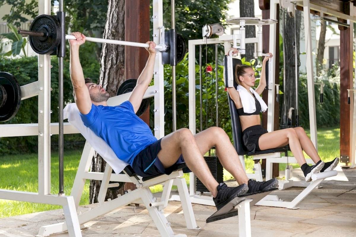 Ein Mann und eine Frau trainieren im Freien unter einem hölzernen Deck mit Fitnessgeräten.