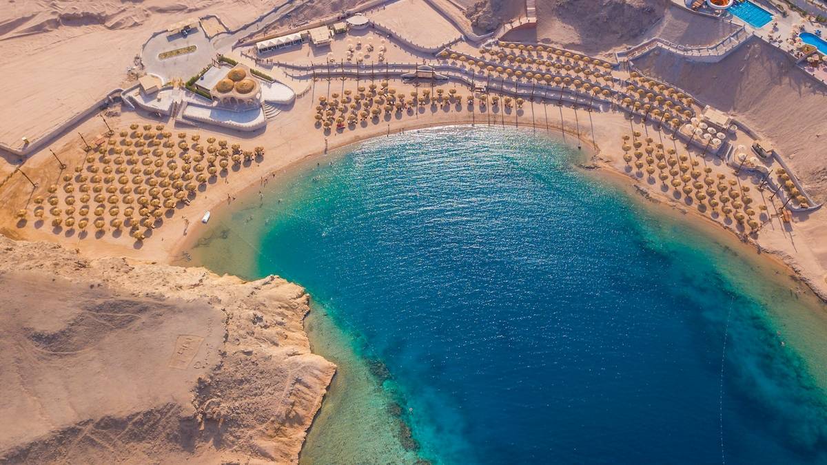 Luftaufnahme des Siva Golden Bay Makadi Hotels, zeigt eine lange Strandlinie mit vielen Sonnenschirmen und einem blauen Meer.