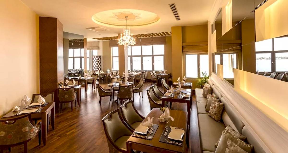 Innenansicht des Restaurants im Melas Resort mit Holzböden, Tischen, Stühlen und einer großen Fensterfront.