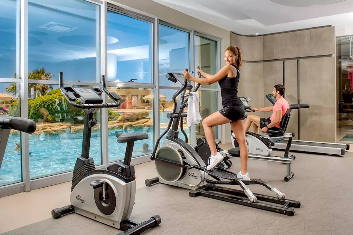 Fitnessraum im Melas Resort mit Frau auf Stepper und Mann auf Laufband neben Swimmingpool.