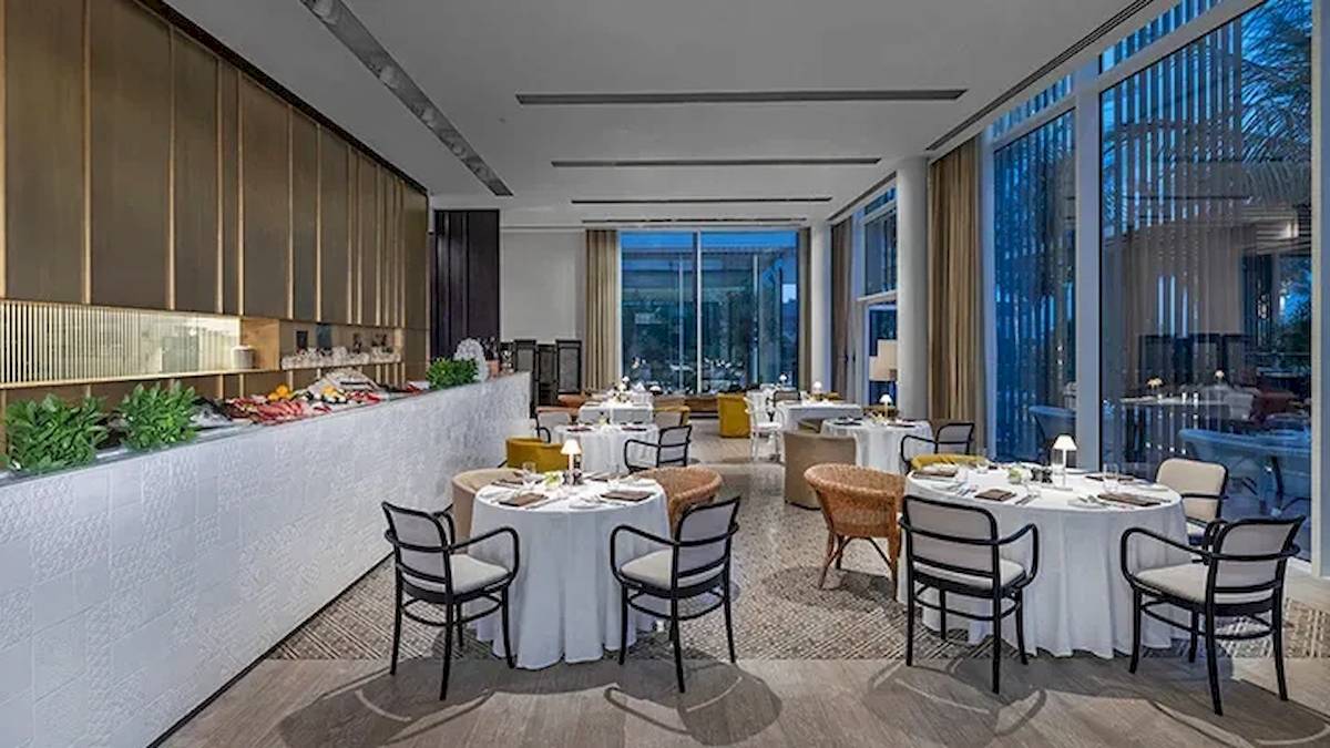 Innenbereich des Restaurants im Oberoi Beach Resort, Al Zorah, mit runden Tischen und Stühlen, großen Fenstern und Pflanzen.