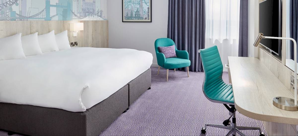 Ein elegantes Hotelzimmer im Leonardo Hotel London Croydon mit einem großen Bett, zwei grünen Stühlen und einem Schreibtisch.