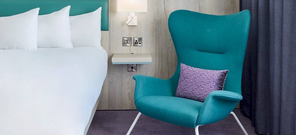 Ein modernes Hotelzimmer mit einem weißen Bett und einem blauen Sessel im Leonardo Hotel London Croydon.