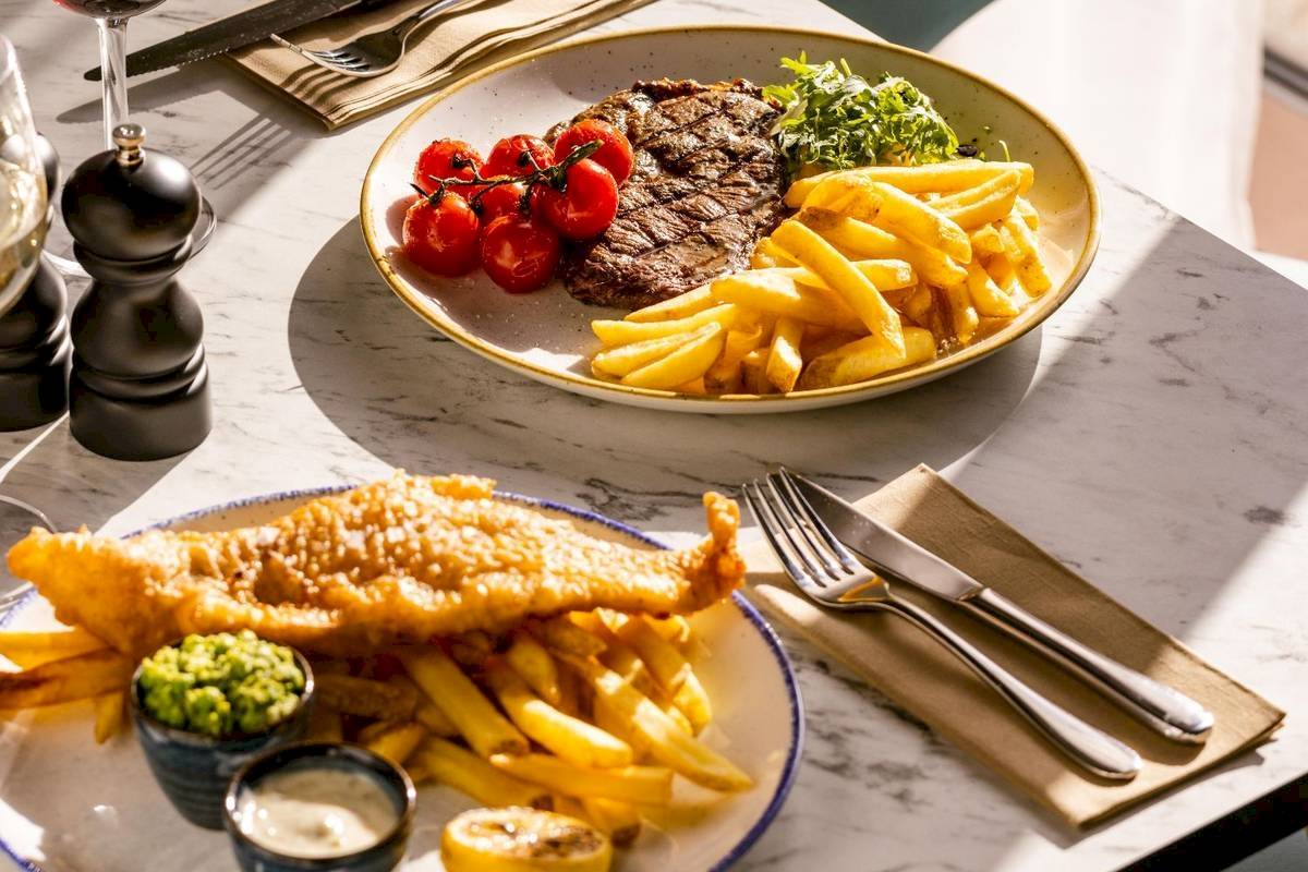 Zwei Teller mit Steak und Pommes sowie Fisch und Pommes auf einem Marmortisch im Leonardo Hotel London Croydon.