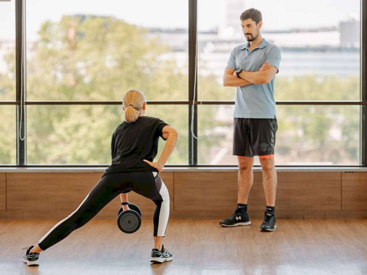 Eine Frau macht eine Übung mit einer Hantel in einem Fitnessstudio, während ein Mann zuschaut.