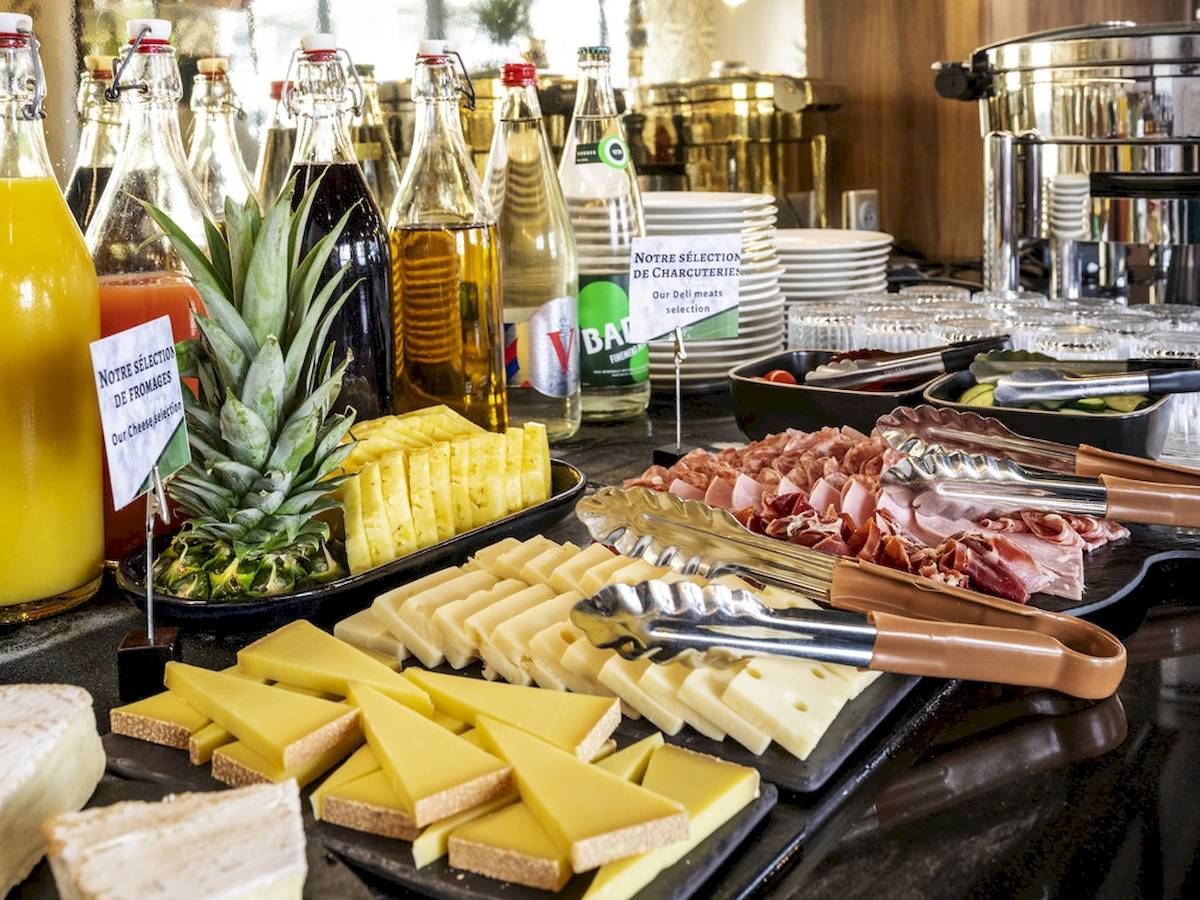 Frühstücksbuffet im Mercure Paris Bercy Bibliothèque mit verschiedenen Käsesorten, Schinken, Tomaten, Orangensaft und weiteren Getränken.