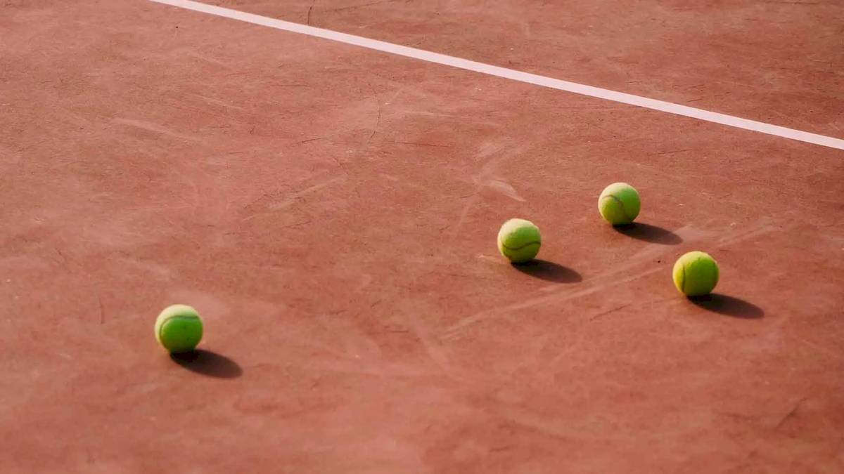 Vier Tennisbälle auf einem roten Tennisplatz neben einer weißen Linie.