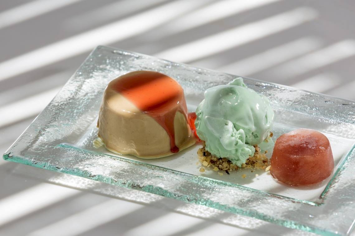 Drei Desserts auf einer Glasplatte, darunter eine Karamellcreme, eine grüne Eiskugel und ein rosa Eiswürfel.