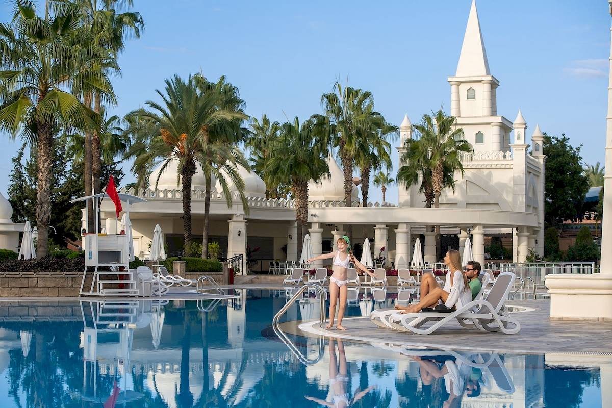 Familie im Sentido Kamelya Fulya Hotel am Pool, mit Palmen und architektonischen Gebäuden im Hintergrund.