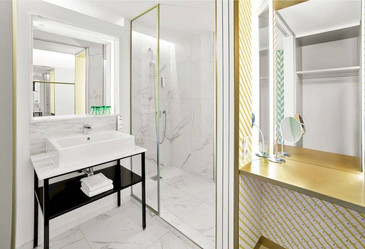 Das elegante Badezimmer des MAXX by Steigenberger Vienna mit weißen Marmorfliesen, goldenen Akzenten und einer modernen Dusche.