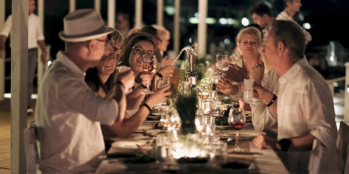 Gäste des Club Marvy sitzen bei einem Abendessen an einem langen Tisch, sie trinken und lachen.