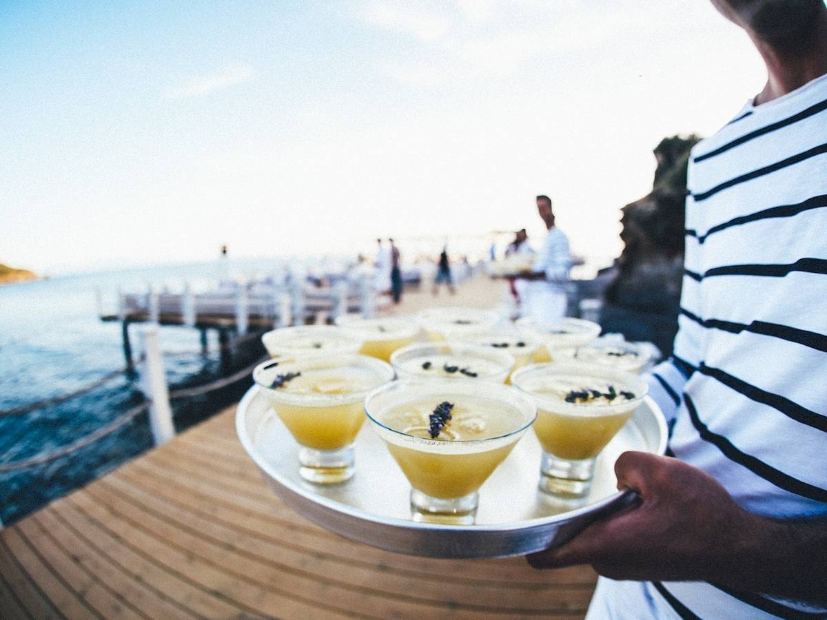 Cocktails auf einem Tablett am Strand, vor einem Hotel im Hintergrund.