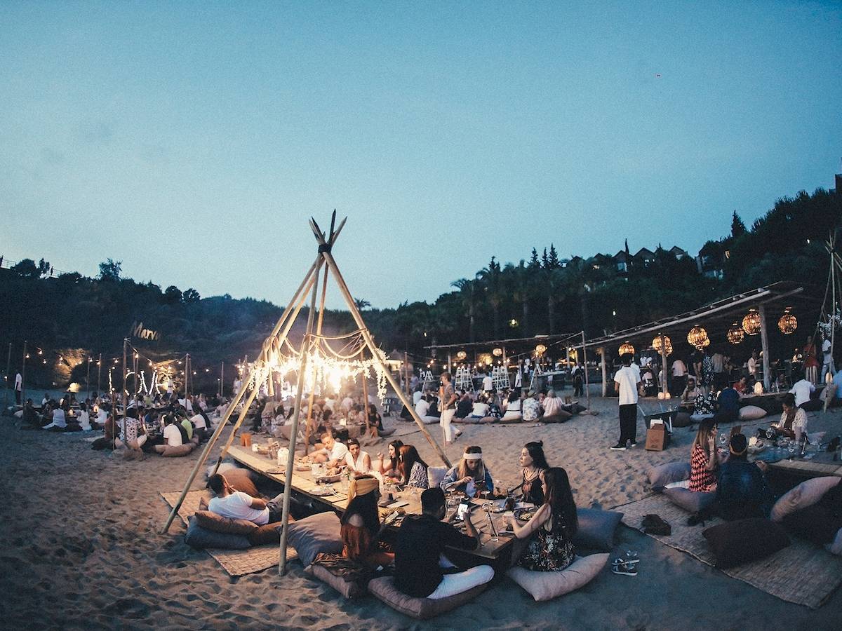 Eine Gruppe von Menschen, die auf Kissen am Strand sitzen und essen, mit einem großen Tipi und Lichterketten im Hintergrund.