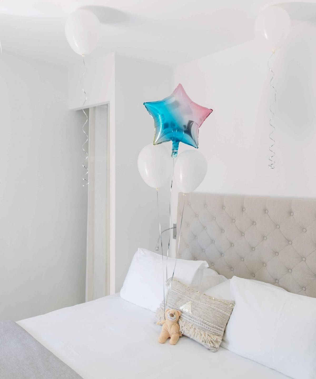 Ein Bett mit weißen Kissen und Ballons, darunter ein Teddybär auf einem gemusterten Kissen.