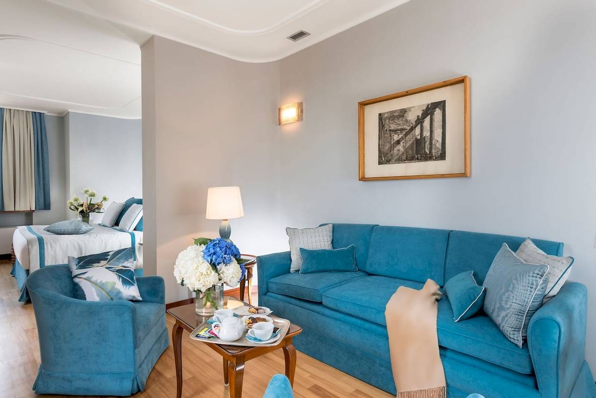 Innenansicht der Suite im Bettoja Hotel Mediterraneo mit blauem Sofa, Tisch, Blumenvase und Bild an der Wand.