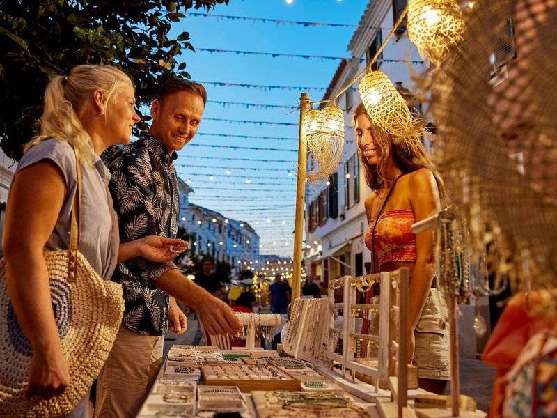 Drei Personen betrachten Schmuck auf einem Marktstand im Abendlicht von Victoria Menorca.