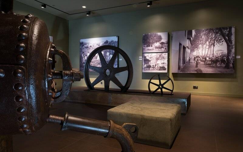 Museumsausstellung mit einem großen Rad und mehreren alten Schwarz-Weiß-Fotos an der Wand.