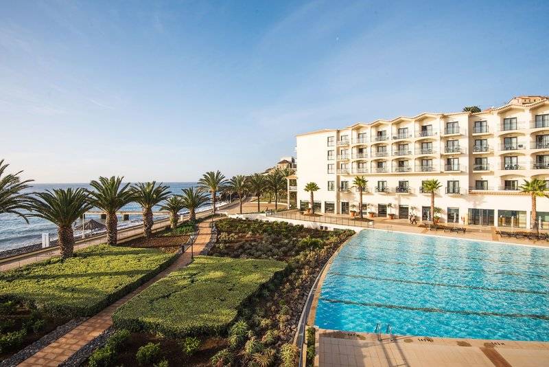 Außenansicht des Vila Galé Santa Cruz Hotels mit Swimmingpool, Palmen und Meer im Hintergrund.
