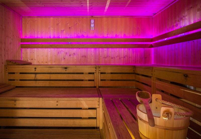 Eine Sauna mit Holzbänken und rosa Beleuchtung, ein Eimer mit einem Holzlöffel daneben.