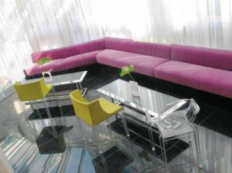 Hotelloungebereich mit einem großen pinken Sofa und gelben und durchsichtigen Stühlen vor einem Glastisch.