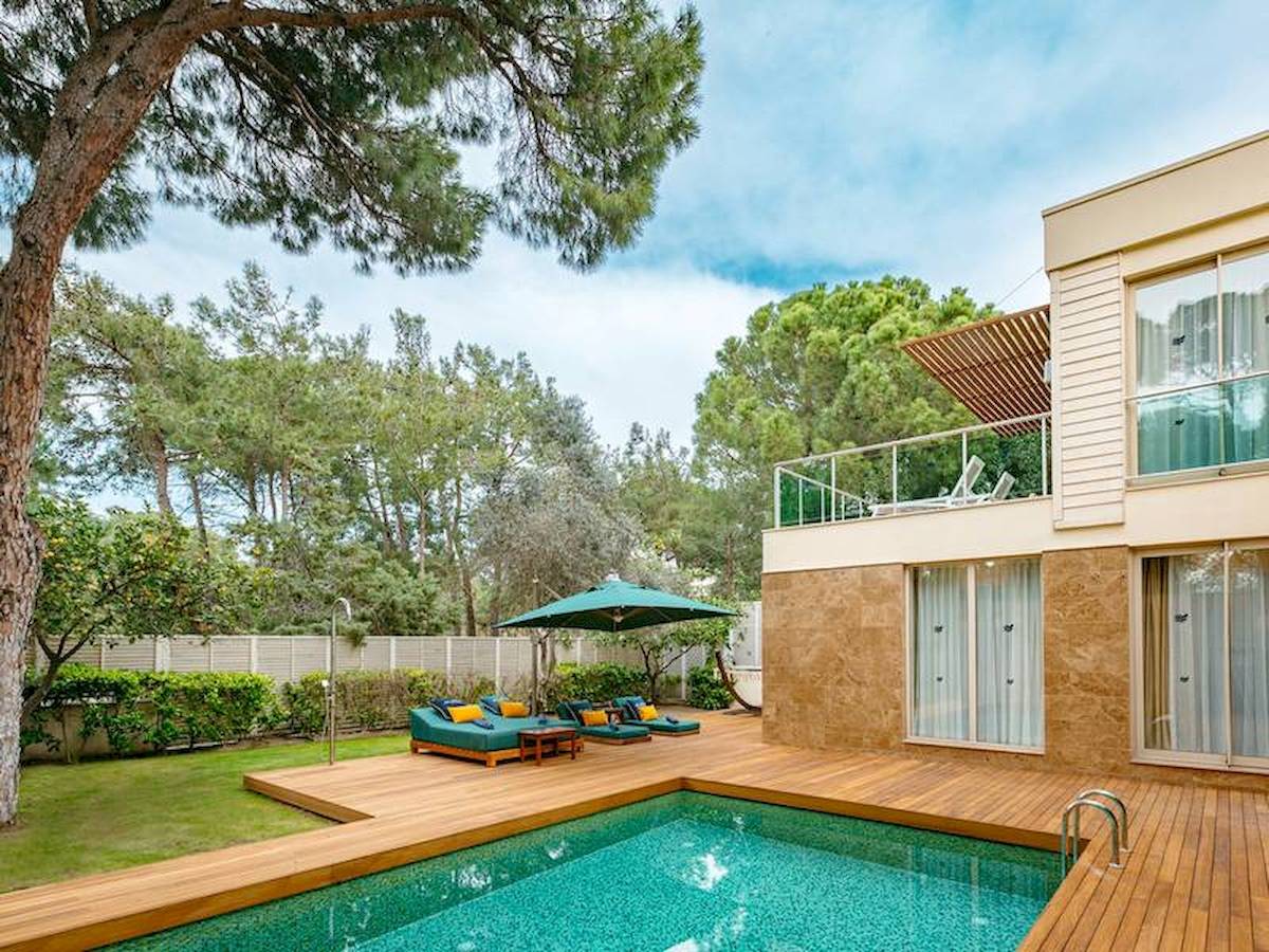 Poolseite der Rixos Premium Belek Villa mit Holzdeck, Loungesofas und einem grünen Sonnenschirm.