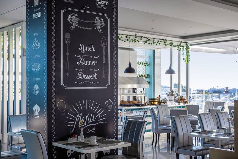 Innenbereich des Asterias Beach Resort Restaurants mit einem großen schwarzen Menüboard, das neben einem Tisch steht, mit Blick auf einen sonnigen Hafen.