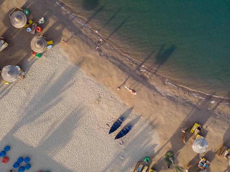 Luftaufnahme des Desert Rose Resorts Strands mit mehreren Liegen, Sonnenschirmen und blauen Booten im Sand.