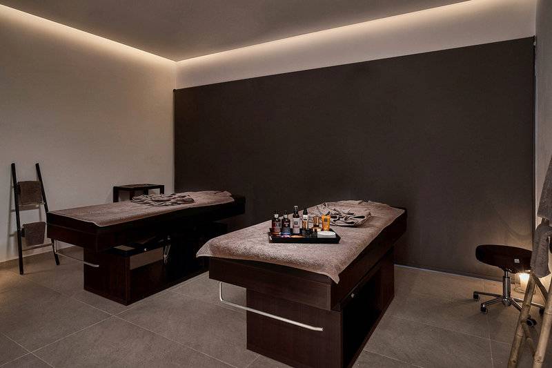 Massageraum im Lindos Grand Resort & Spa mit zwei Massagetischen und einem Stuhl.