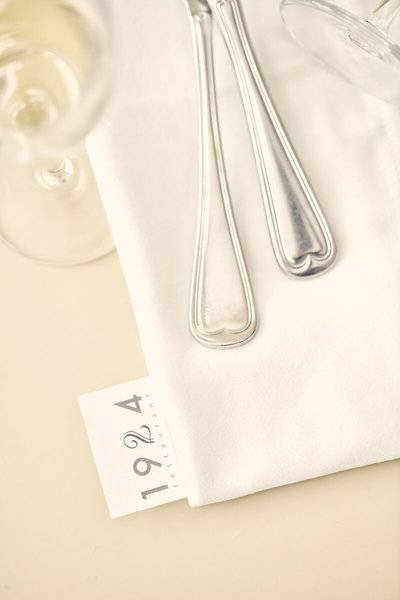 Ein Tisch im Lagos Mare Hotel, elegant eingedeckt mit einem weißen Tuch, Silberbesteck und einer Platzkarte mit dem Jahr 1924.
