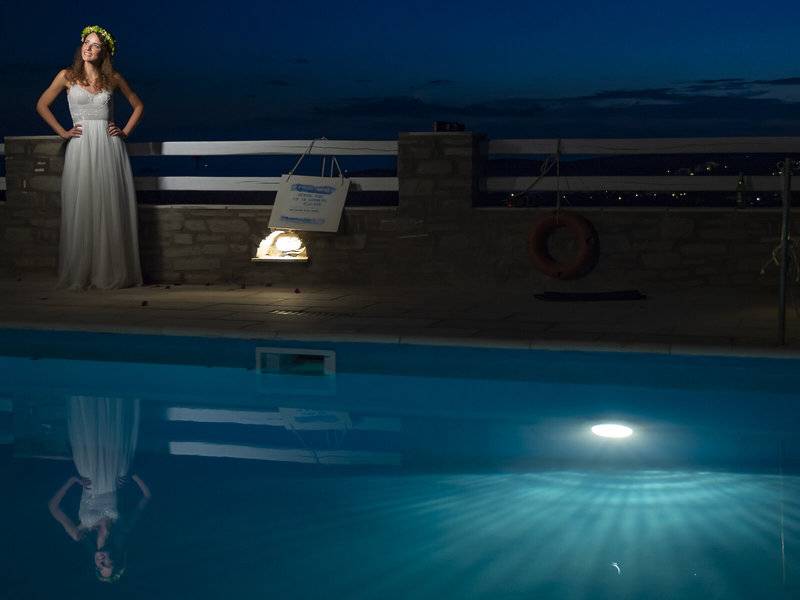 Eine Frau im weißen Kleid steht am Rand eines Swimmingpools bei Nacht.