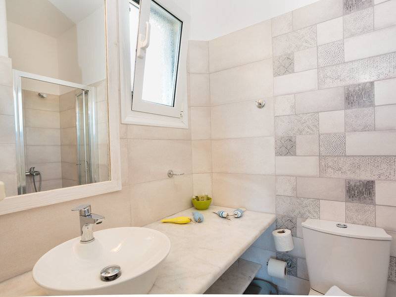 Badezimmer im Pyrgaki Hotel mit weißem Waschbecken, Spiegel, Duschkabine und gefliester Wand.