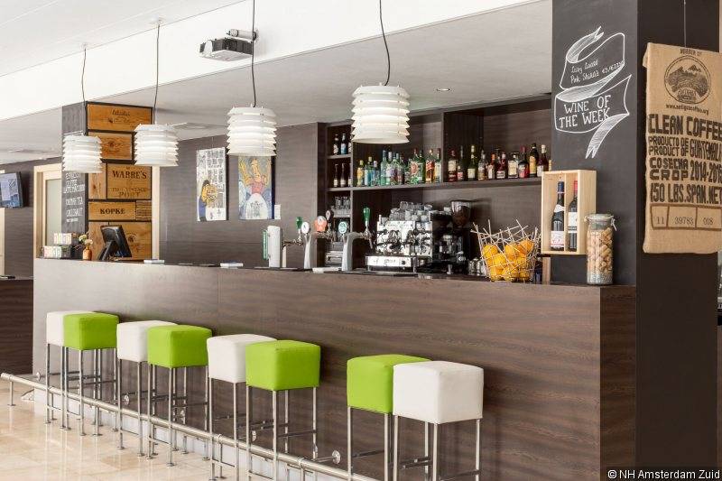 Hotelbar des NH Amsterdam Zuid mit grünen und weißen Hockern, Theke, Getränkespendern und Barkeeperstation.