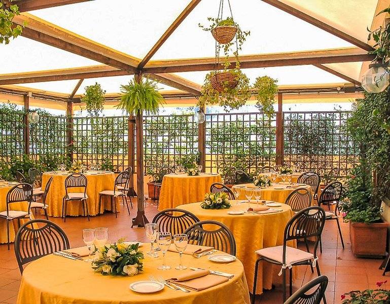Garten-Terrasse des Pineta Palace Hotels mit runden Tischen, Stühlen und gelben Tischdecken, umgeben von Pflanzen und Blumen.