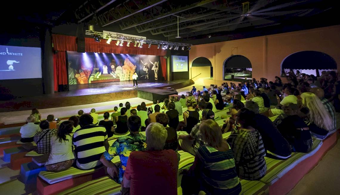 Ein Publikum sitzt in einem Theatersaal und schaut eine Live-Show auf der Bühne, während ein Mann in schwarzer Kleidung auf der Bühne steht, während es Nacht ist.