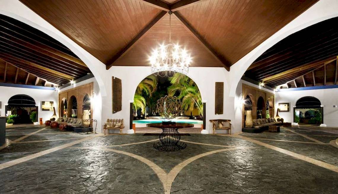 Innenansicht des Hotel-Lobbys des Catalonia Bayahibe mit einer großen Deckenleuchte, einem zentralen Tisch, einem Swimmingpool im Hintergrund und Möbeln.