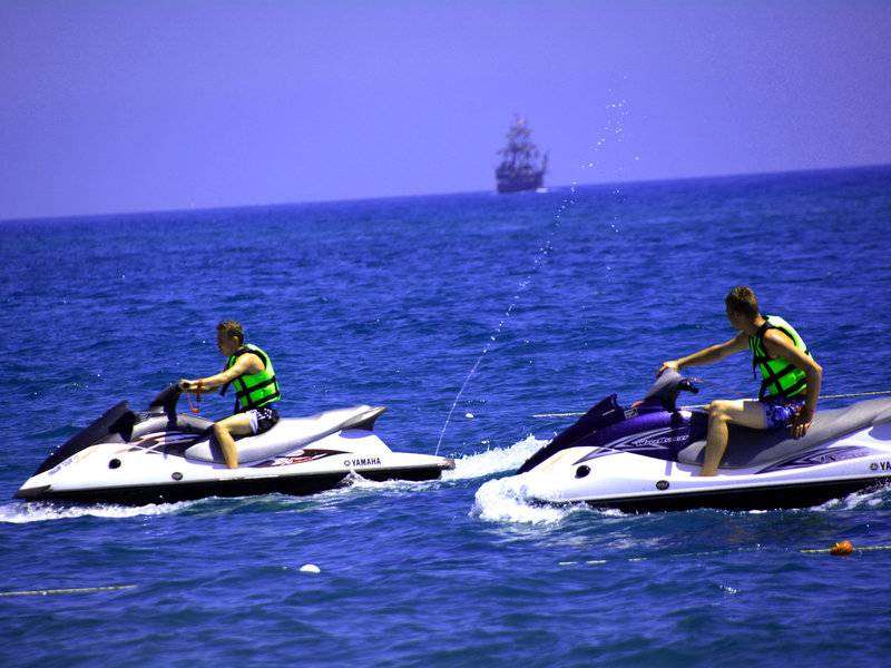 Zwei Personen auf Jetskis im Meer, ein Segelschiff im Hintergrund.