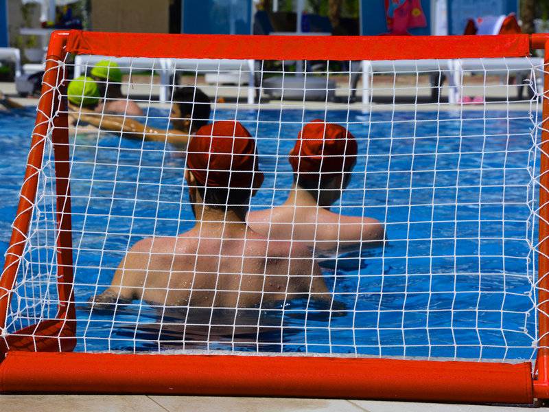 Schwimmer im Schwimmbad mit roten Hüten spielen Wasserball vor einem roten Tor.