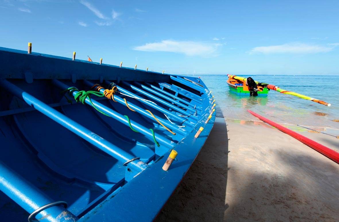 Ein blaues Boot am Strand mit einem Ruderboot im Wasser an einem sonnigen Tag.