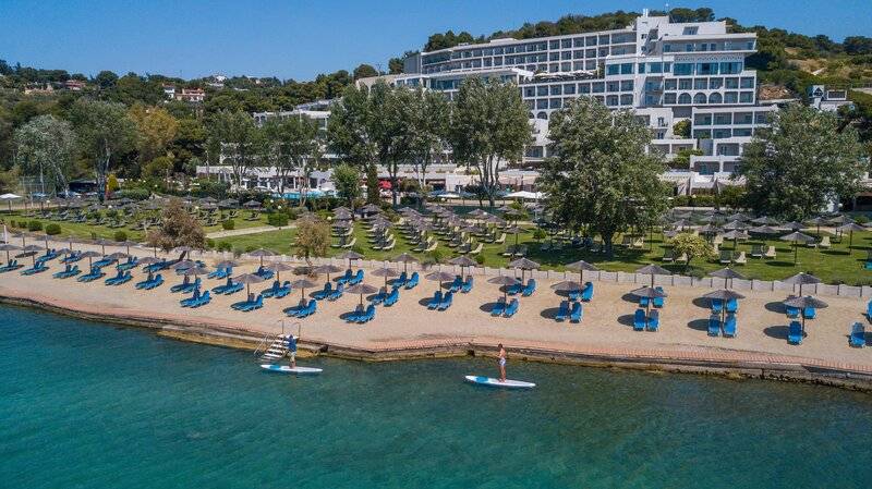 Luftaufnahme des Dolce Athens Attica Riviera Hotels mit vielen Sonnenliegen und Sonnenschirmen am Strand