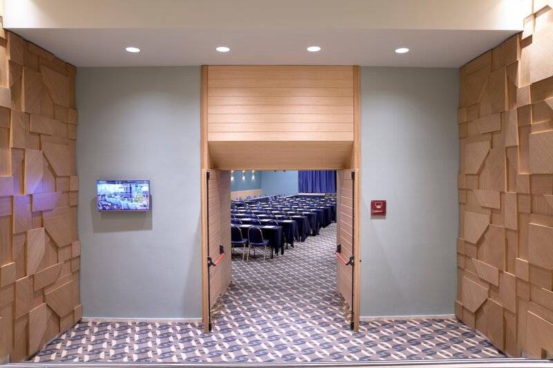 Eingangsbereich des Konferenzraums im Hotel Dolce Athens Attica Riviera mit geometrischen Holzwandverkleidungen und einer Fernsehwand
