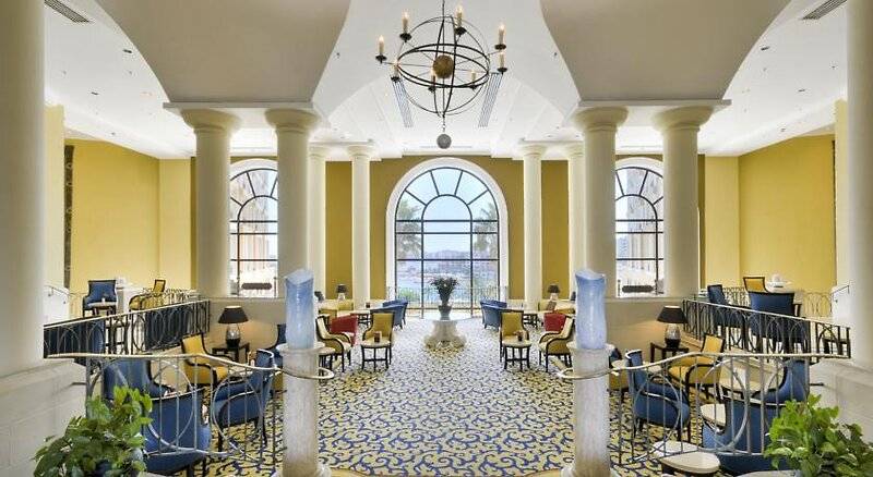 Lobby des Corinthia St. George's Bay Hotels mit gelben Wänden, Säulen und blauen Stühlen.