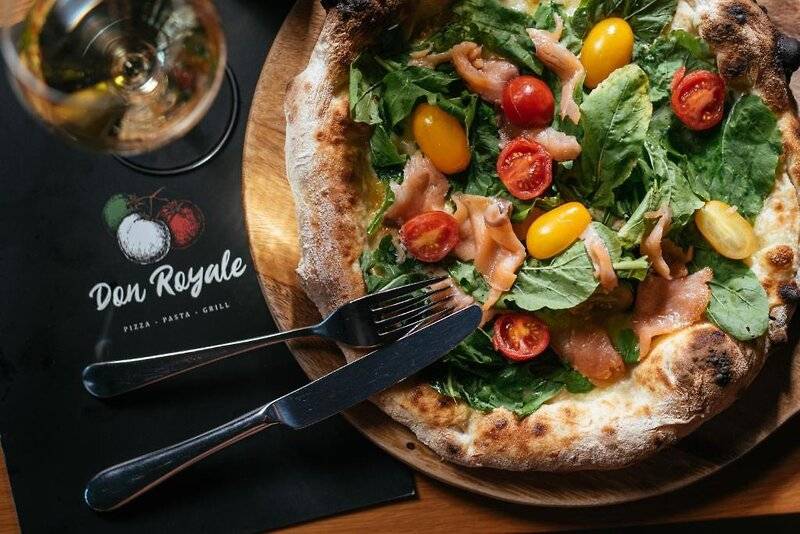 Eine Pizza mit Lachs und Tomaten auf einer Holzplatte mit Messer und Gabel.