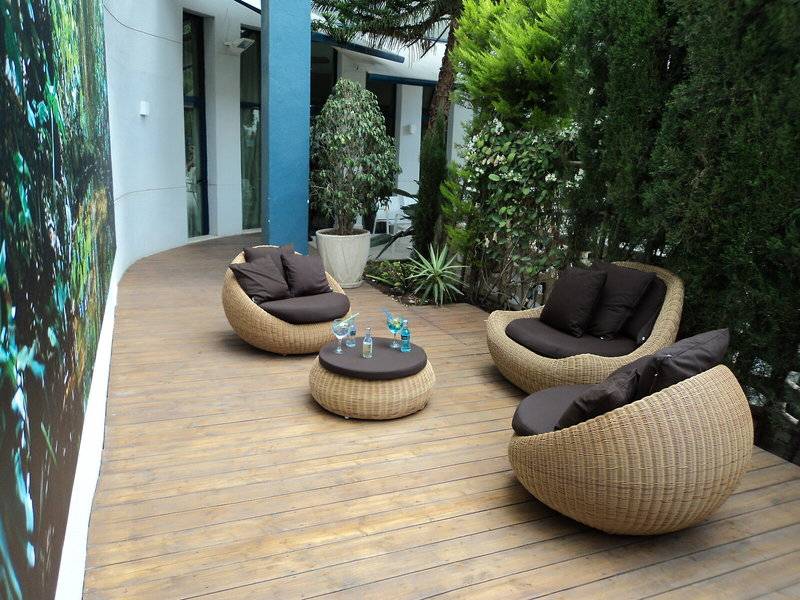 Terrasse mit Korbmöbeln und einem Tisch im Freien, umgeben von Pflanzen und einer Wandmalerei.