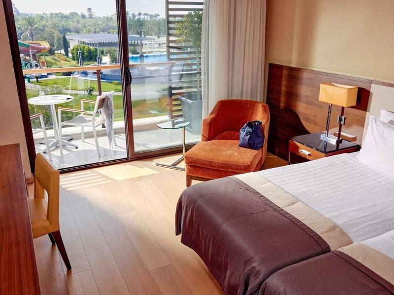 Doppelzimmer im TUI BLUE Side Hotel mit Balkon, zwei Betten, Tisch, Stuhl und Aussicht auf Pool und Rutsche.