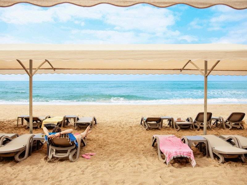 Person entspannt sich auf einem Liegestuhl unter einem Sonnenschirm am Strand des Magic Life Jacaranda Hotels.