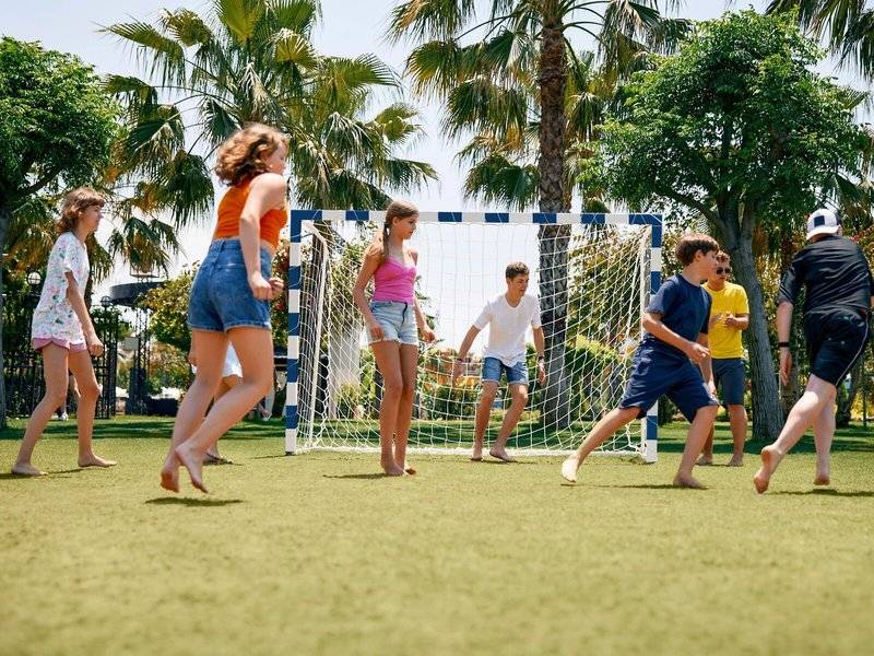 Mehrere Kinder spielen Fussball auf einem Kunstrasenplatz im Freien, vor einem Tor.