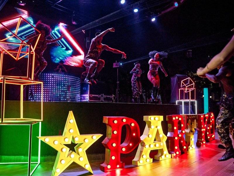 Party auf der Bühne im Magic Life Jacaranda Hotel mit leuchtenden Neon-Lettern und tanzenden Gästen.